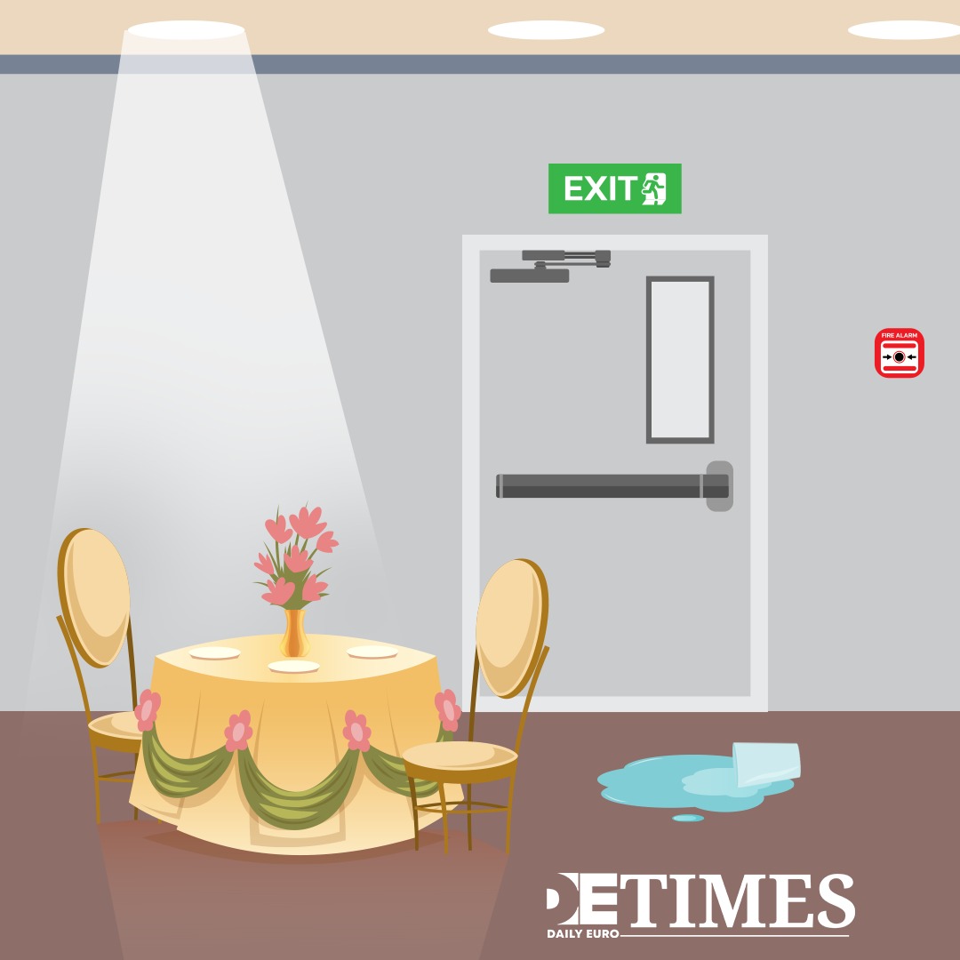 Round dining table set for four under a spotlight, with a pink flower centerpiece, in a banquet room beside an exit door.